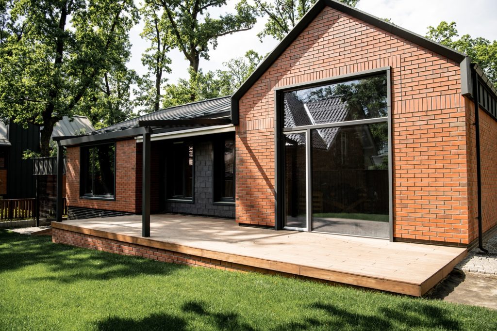 Dom z ceglaną elewacją, dużymi oknami aluminiowymi i drewnianym tarasem w otoczeniu zieleni